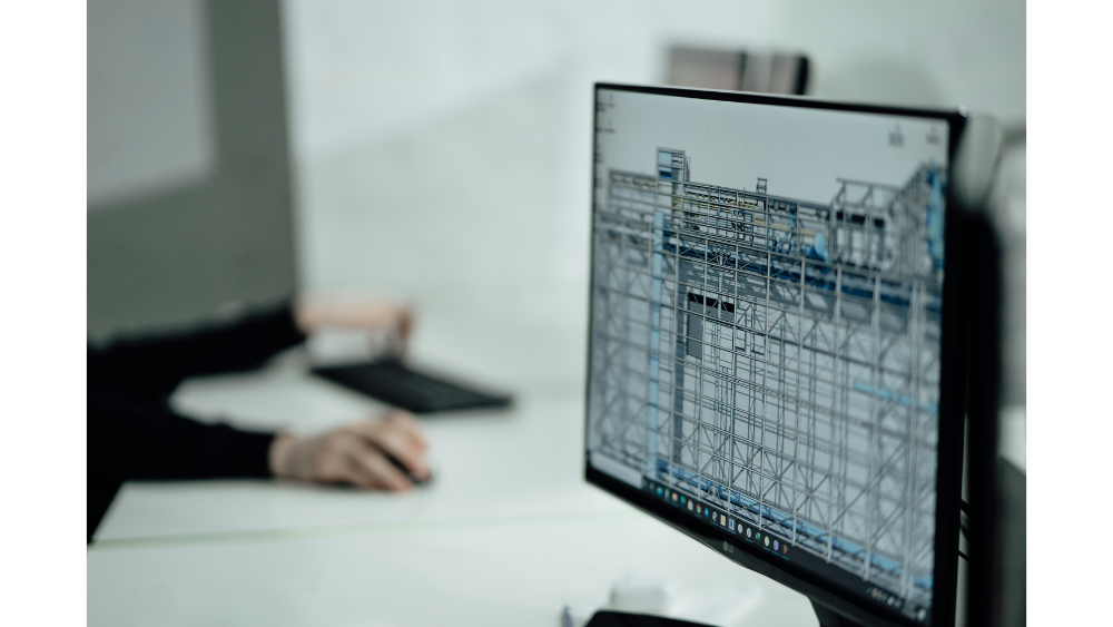 A computer monitor showing a digital wireframe model of a building while a person works in the background using a keyboard and mouse.
